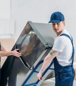 two-movers-in-uniform-using-hand-truck-while-transporting-refrigerator-in-apartment